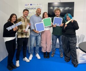 Team BT Group in piedi nello stand BT Group a Didacta 2025, su pavimento blu, con cartelli colorati e scritte “TOOLBOX” in mano. Sullo sfondo un monitor nero e un pannello informativo con il logo TOOLBOX.