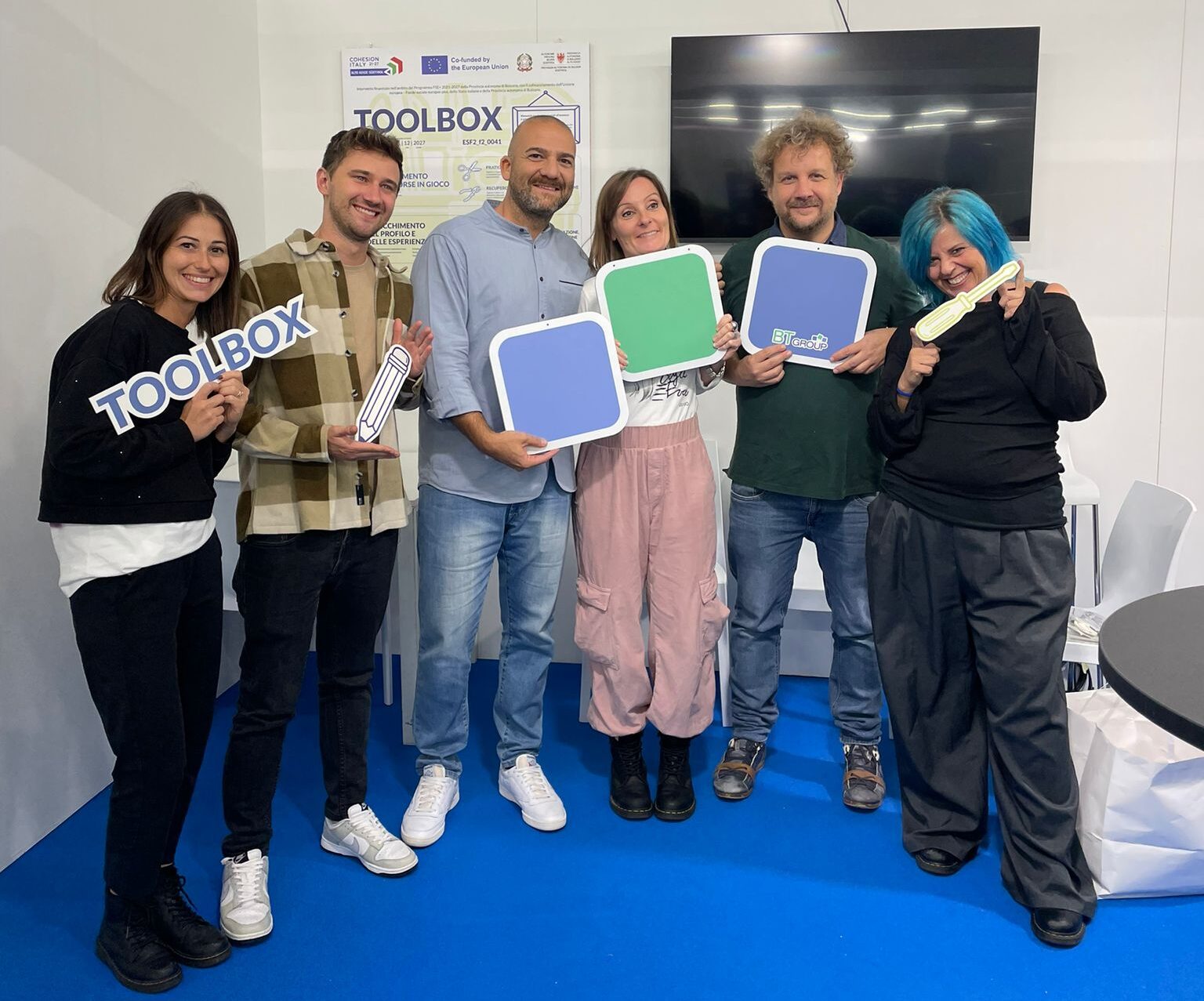 Team BT Group in piedi nello stand BT Group a Didacta 2025, su pavimento blu, con cartelli colorati e scritte “TOOLBOX” in mano. Sullo sfondo un monitor nero e un pannello informativo con il logo TOOLBOX.
