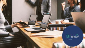 Persone in riunione aziendale attorno a un tavolo con laptop e documenti, mentre un collega presenta davanti a una lavagna. BT Voices: le imprese raccontano le competenze del futuro nei nuovi talenti.