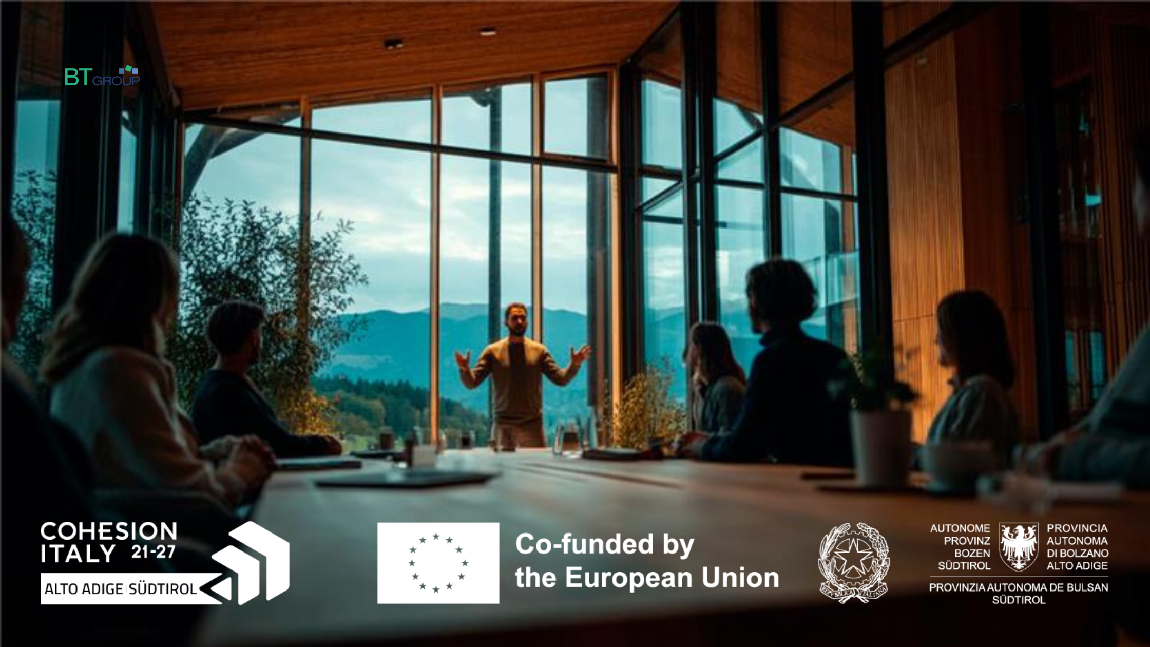 Persone sedute attorno a un tavolo in una sala riunioni luminosa con ampie vetrate, vista sulle montagne dell’Alto Adige. Un relatore presenta davanti al gruppo. Immagine dedicata alla formazione continua in Provincia di Bolzano, btgroup.tn.it.