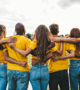 Un gruppo di giovani, uniti fianco a fianco con le braccia sulle spalle, indossa magliette gialle mentre guarda verso l’orizzonte in uno spazio all’aperto. La scena rappresenta inclusione, cooperazione e protagonismo giovanile, temi centrali del progetto ITER per la riattivazione, la riconnessione e la rigenerazione delle comunità.